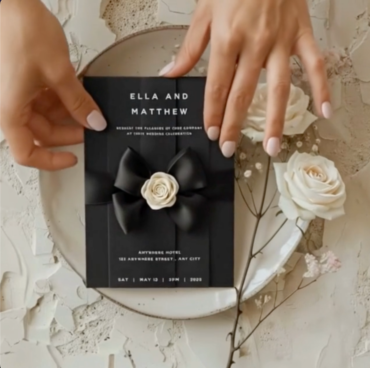 Hands placing a black and white wedding invitation with a satin bow and camellia flower on a plate, surrounded by white roses and soft neutral textures minimalist luxury wedding styling video.